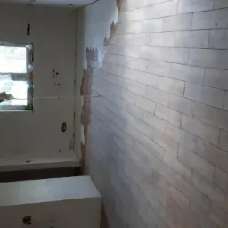 Wnętrze w trakcie remontu, widoczna ułożona podłoga z paneli imitujących drewno, ściany przygotowane do malowania, okno z widokiem na zewnątrz.