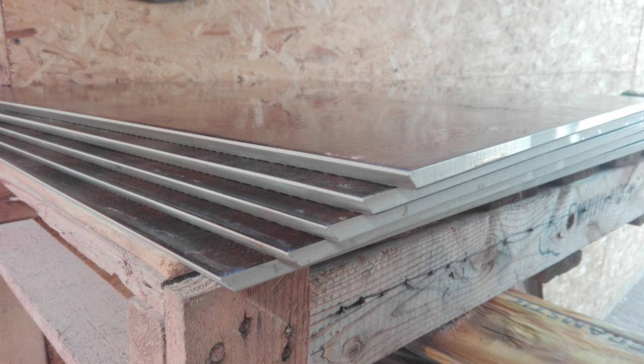 Stos płyt gipsowo-kartonowych z widoczną strukturą warstwową, ułożonych na drewnianej palecie w magazynie budowlanym.