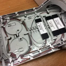 Otwarta skrzynka telekomunikacyjna z uporządkowanymi kablami światłowodowymi i trzema modułami oznaczonymi SPR 08, SPR 09 i SPR 10, leżąca na drewnianym blacie.