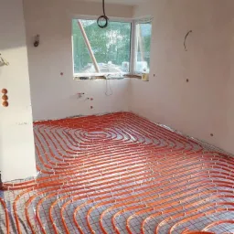 Pomarańczowe rury ogrzewania podłogowego ułożone na metalowej siatce w surowym pomieszczeniu z oknem, widoczne niedokończone ściany i wystające przewody.