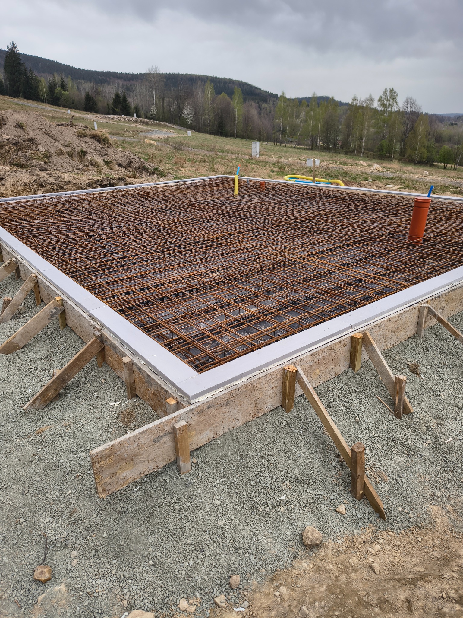 Zbrojenie fundamentów pod budynek jednorodzinny. Widoczne szalunki drewniane, siatka zbrojeniowa i wystające rury instalacyjne. W tle zielony krajobraz i pochmurne niebo.