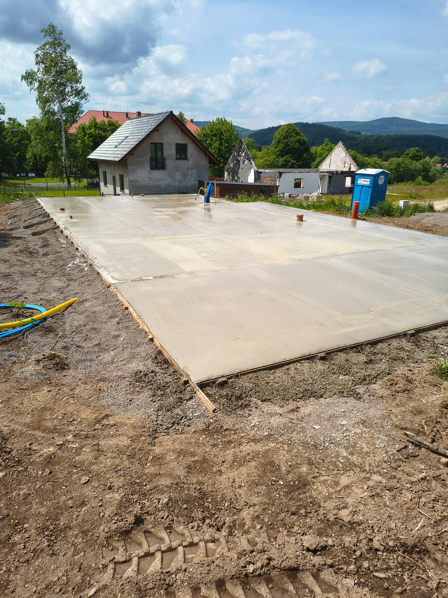 Świeżo wylana płyta fundamentowa pod budynek w Sieradzu. Widoczne szalunki i instalacje. W tle dom w stanie surowym i przenośna toaleta.