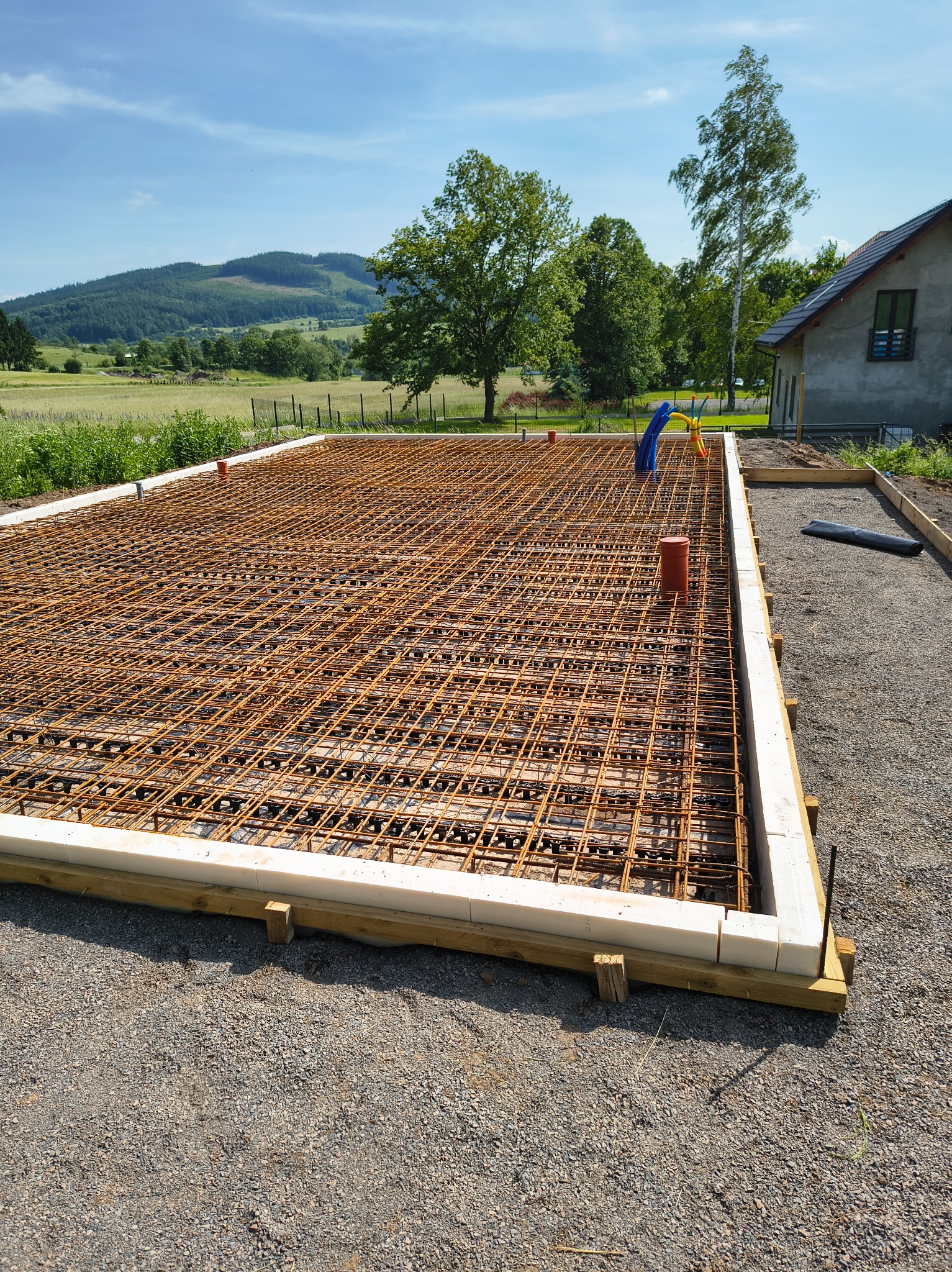 Zbrojenie fundamentu domu jednorodzinnego z widocznymi elementami instalacji, wykonane na zewnątrz w słoneczny dzień z widokiem na malowniczy krajobraz.