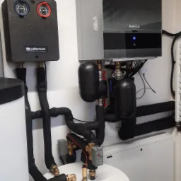 Nowoczesny piec gazowy Buderus z podłączeniami, widoczne zawory, rury z izolacją termiczną i zbiornik na wodę, zamontowany na białej ścianie.