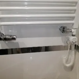 Biały, drabinkowy grzejnik łazienkowy z chromowanymi zaworami i elektrycznym sterownikiem zasilanym spiralnym przewodem.