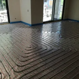 Rozłożone rury ogrzewania podłogowego na izolacji z czerwonym rastrem, zabezpieczone niebieską folią przy ścianach w surowym wnętrzu z dużymi oknami.