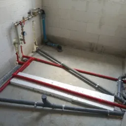 Instalacja wodno-kanalizacyjna i grzewcza w surowym stanie: czerwone rury grzewcze ułożone w ramę, szare rury kanalizacyjne z obejmami, białe płyty izolacyjne na podłodze, widoczne liczniki...