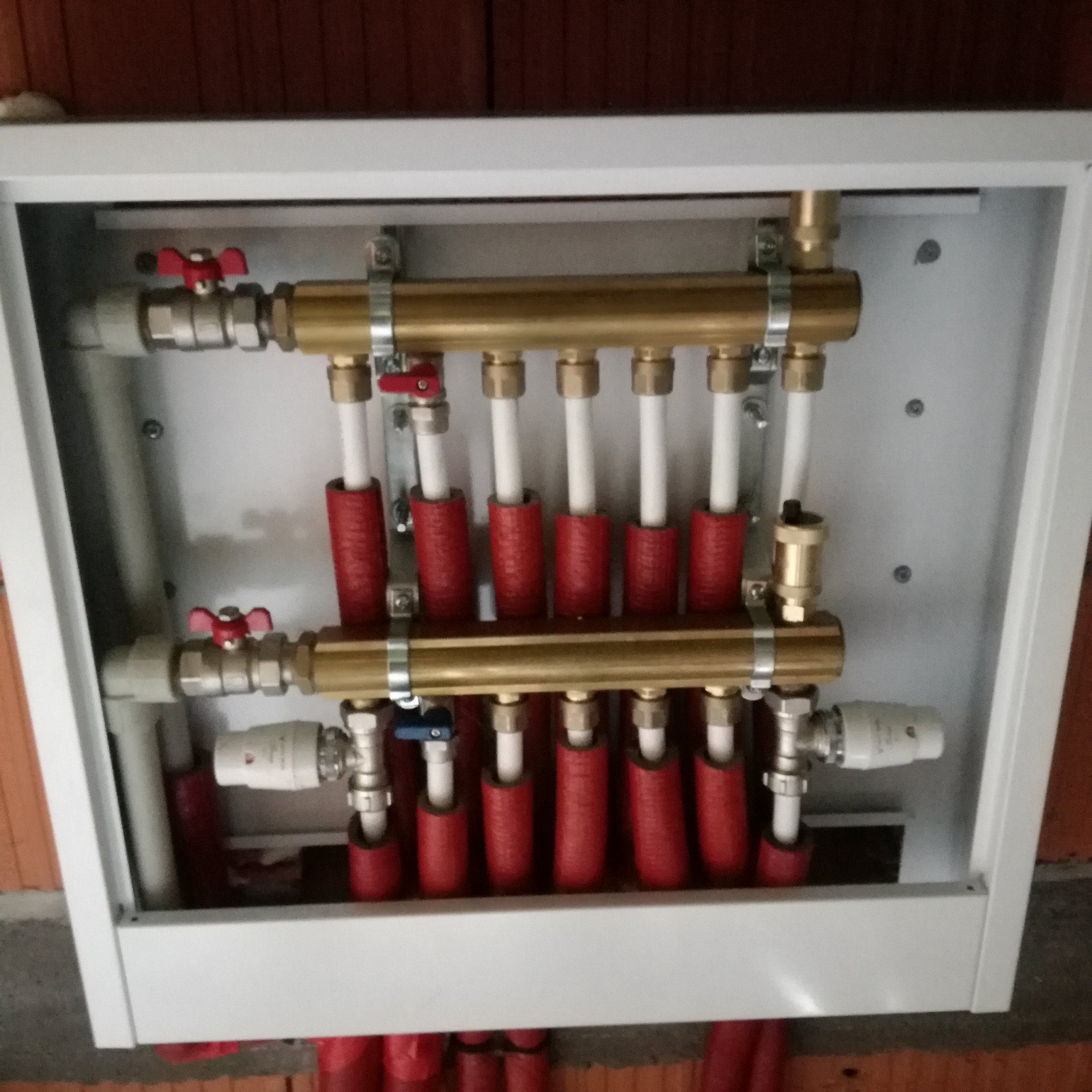 Rozdzielacz ogrzewania podłogowego w białej szafce, widoczne mosiężne rury, białe rurki z czerwonymi otulinami, zawory z czerwonymi pokrętłami, zamontowany na tle ściany z cegły.