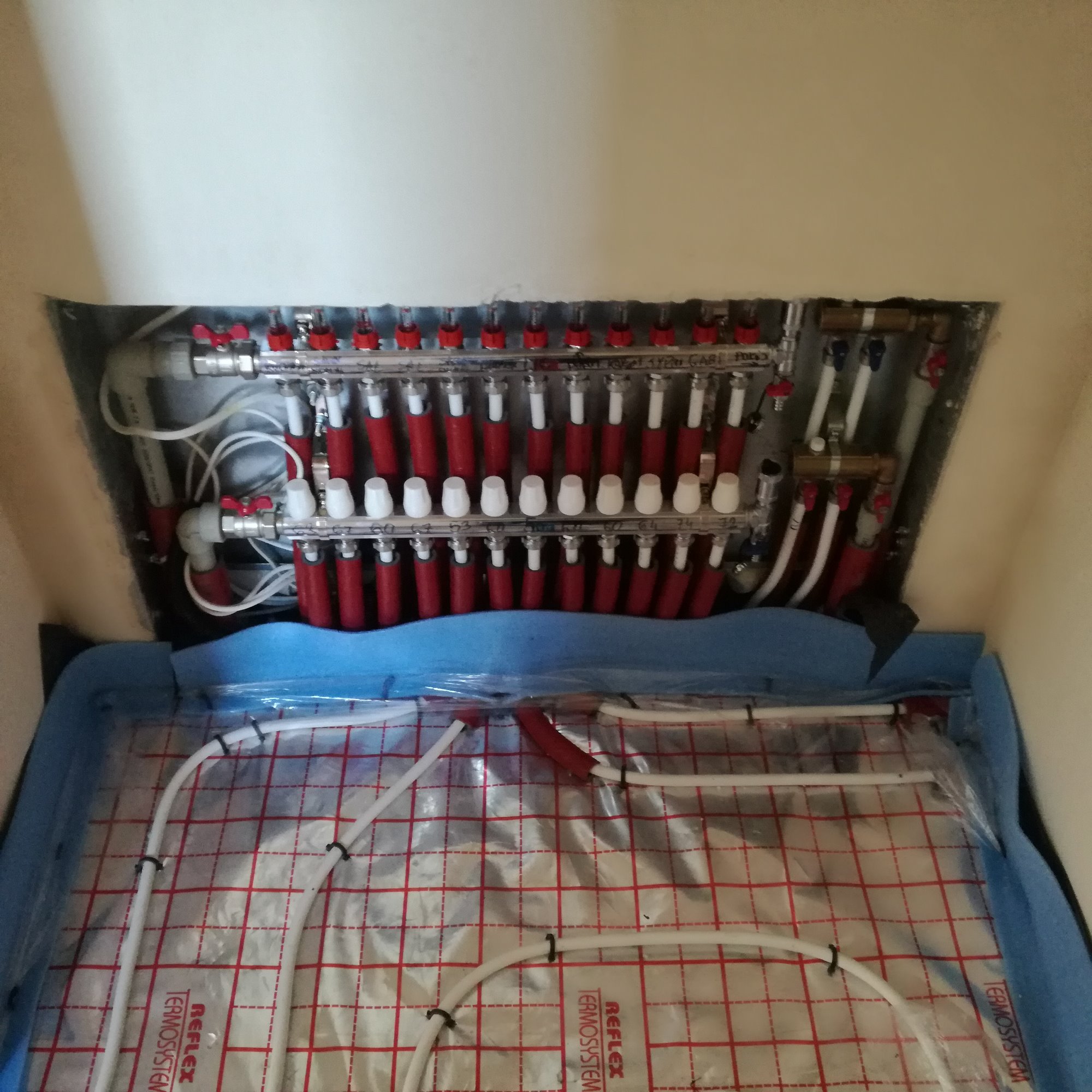 Rozdzielacz ogrzewania podłogowego z czerwonymi zaworami i białymi rurami, widoczne rury ułożone na folii z czerwoną kratką, niebieska izolacja na krawędziach.