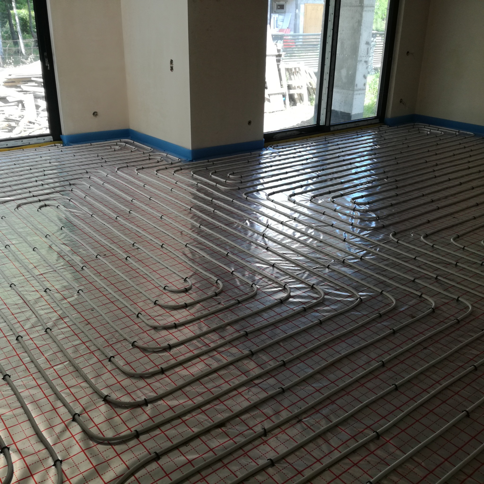 Rozłożone rury ogrzewania podłogowego na izolacji z czerwonym rastrem, zabezpieczone niebieską folią przy ścianach w surowym wnętrzu z dużymi oknami.