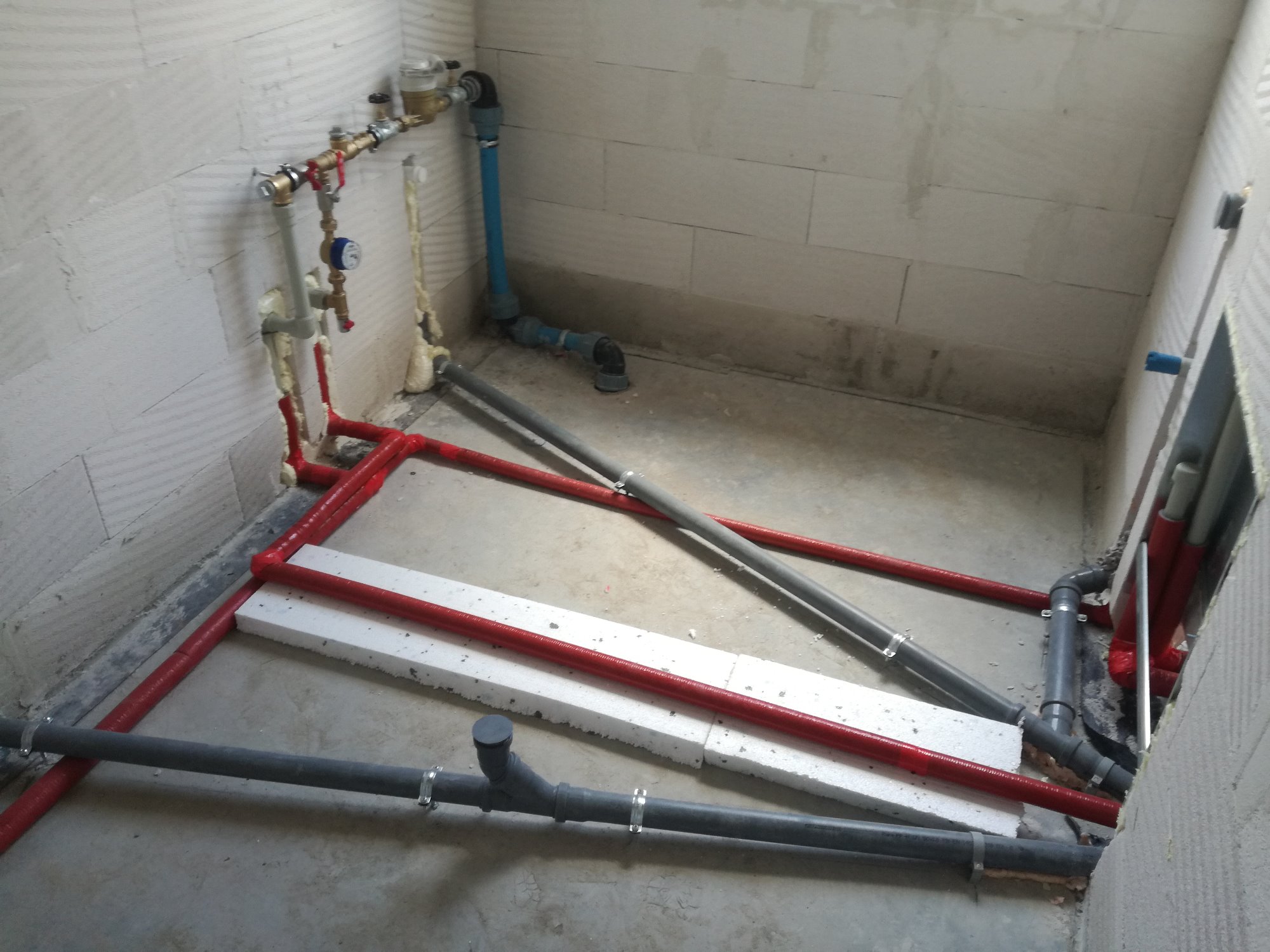 Instalacja wodno-kanalizacyjna i grzewcza w surowym stanie: czerwone rury grzewcze ułożone w ramę, szare rury kanalizacyjne z obejmami, białe płyty izolacyjne na podłodze, widoczne liczniki...