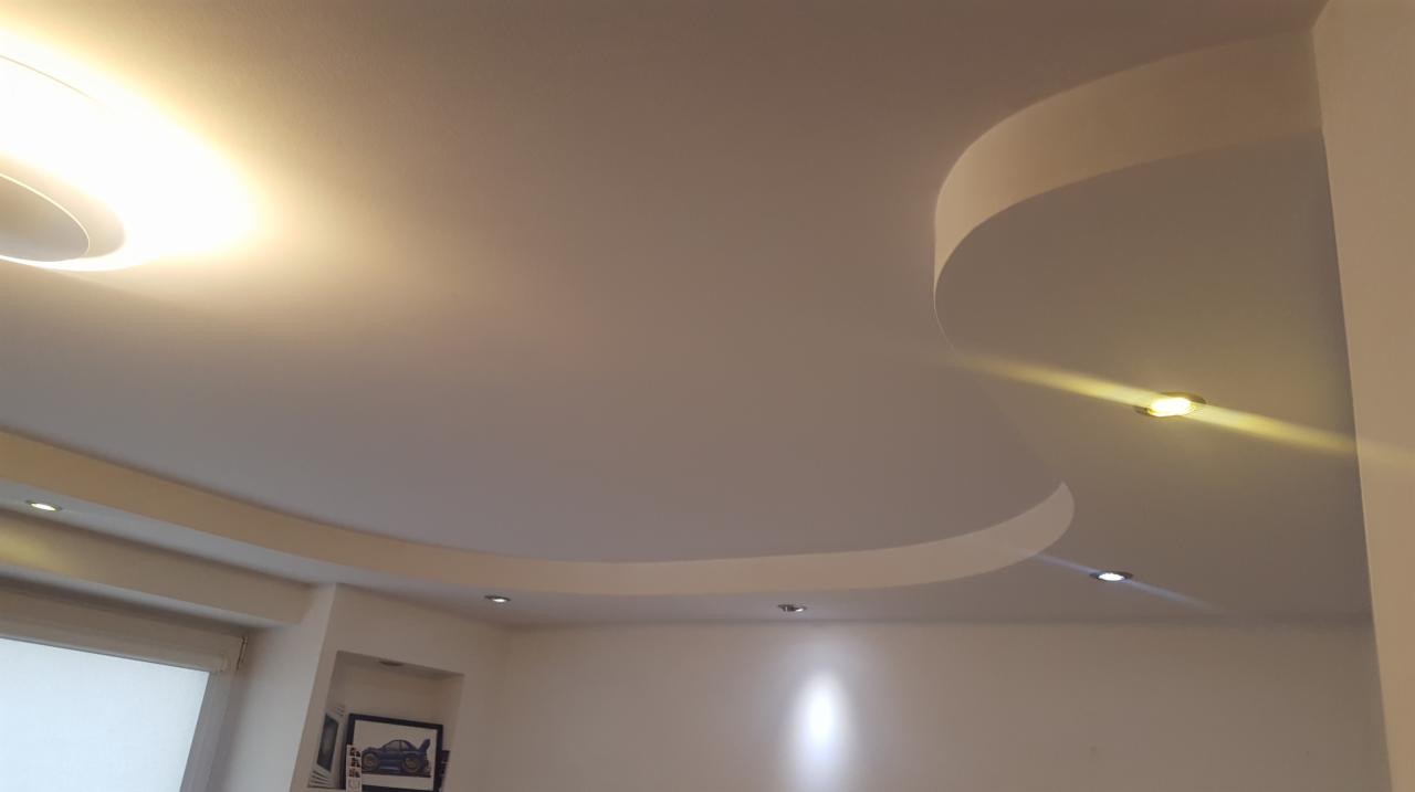 Gładki, biały sufit w nowoczesnym salonie z podwieszanymi elementami w kształcie łuków, oświetleniem punktowym i okrągłą lampą sufitową dającą rozproszone światło.