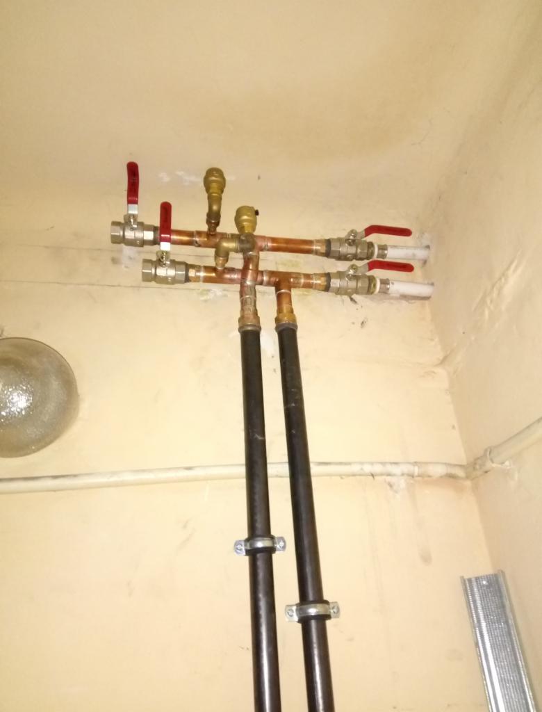 Fragment instalacji hydraulicznej z zaworami z czerwonymi uchwytami, rurami miedzianymi i czarnymi rurami stalowymi przymocowanymi do ściany klamrami. Widoczne łączenia i elementy instalacji.