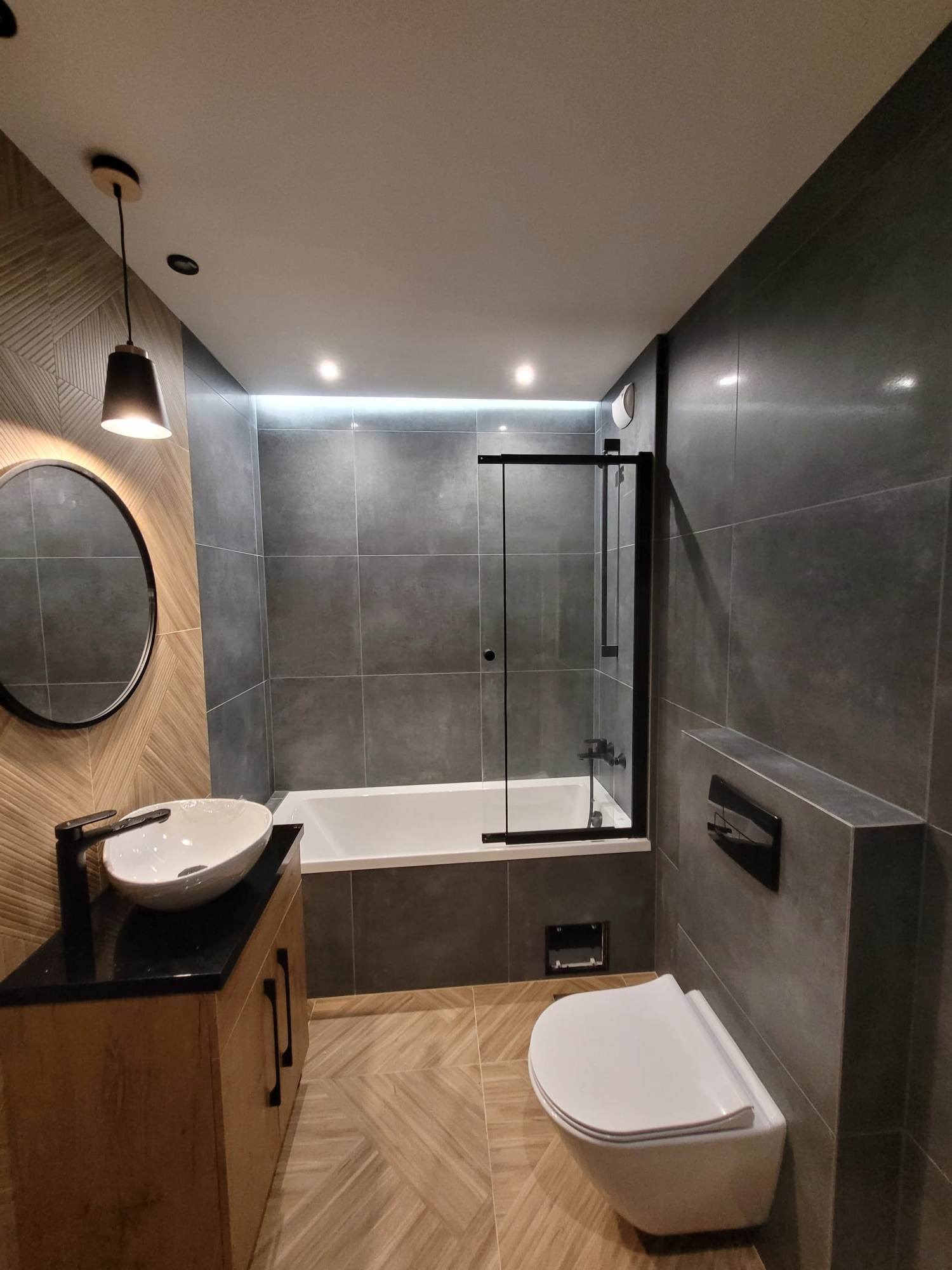 Nowoczesna łazienka z szarymi płytkami, drewnianą szafką pod umywalką z czarnym blatem, okrągłym lustrem i kabiną prysznicową z czarnymi ramami.