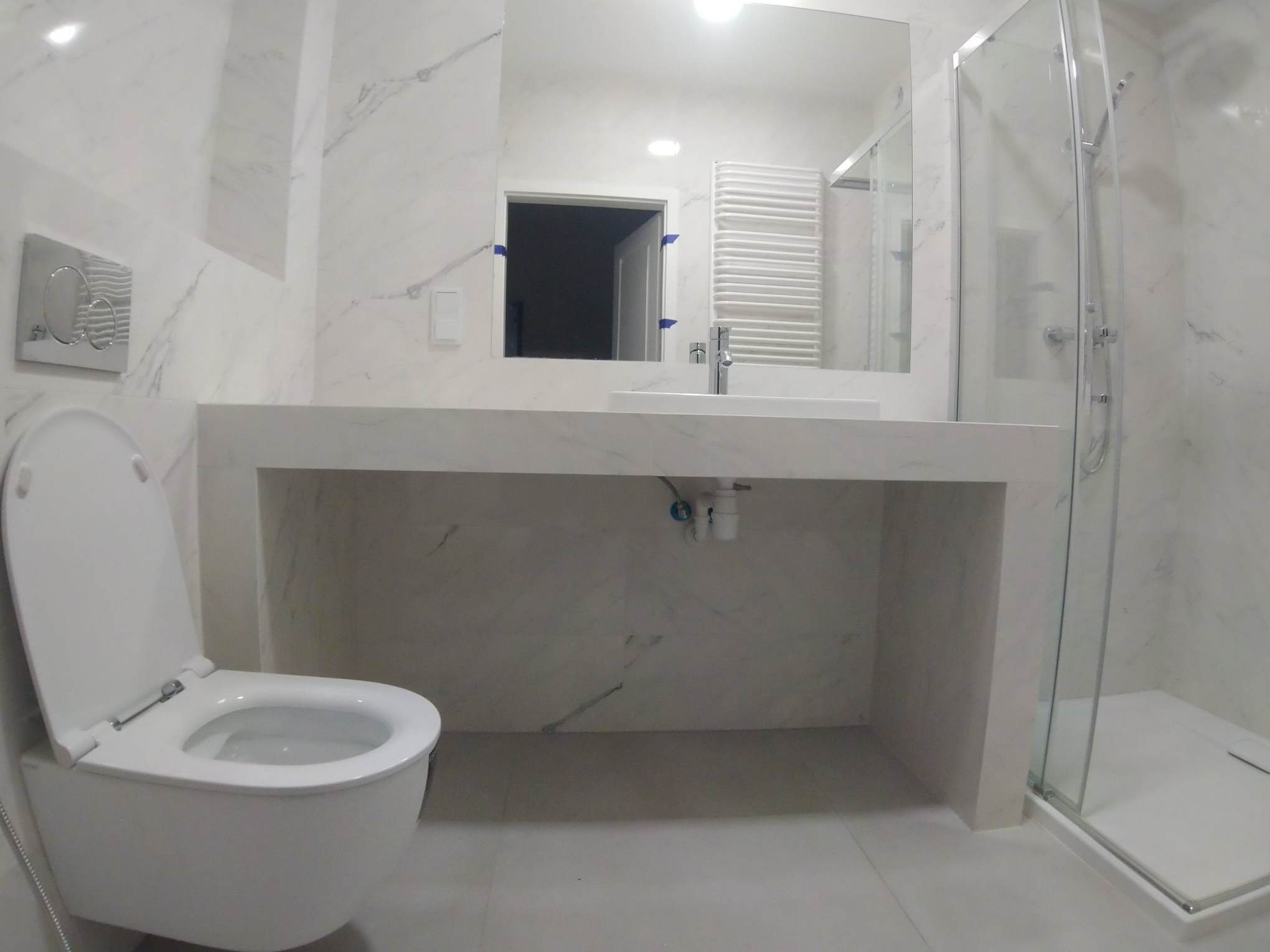 Nowoczesna łazienka w bieli z marmurowym wzorem, wisząca toaleta z otwartą deską, przeszklona kabina prysznicowa i umywalka wpuszczona w blat, minimalistyczny grzejnik drabinkowy.