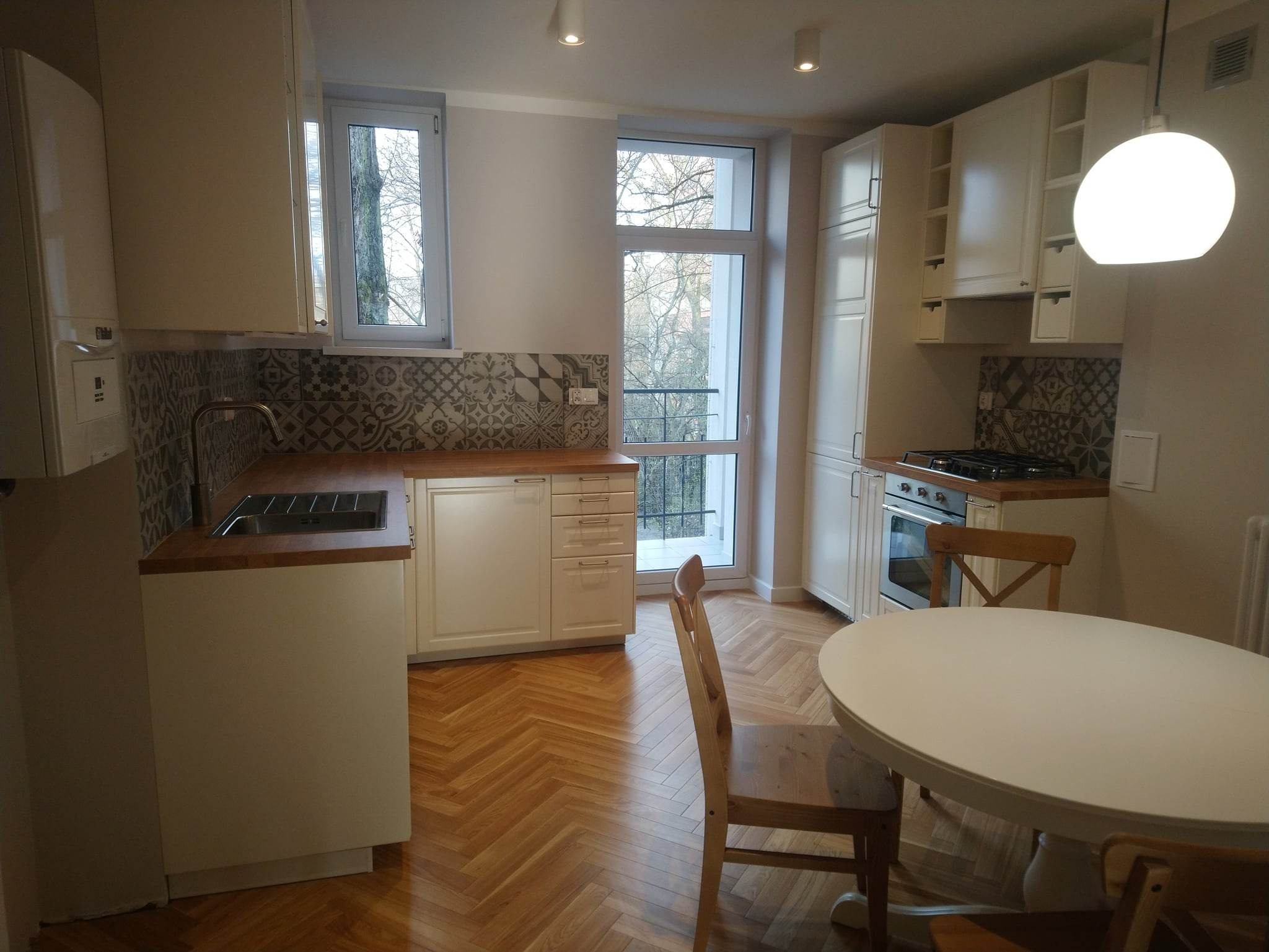 Jasna kuchnia w stylu klasycznym z drewnianym parkietem w jodełkę, okrągłym białym stołem i krzesłami z naturalnego drewna, wzorzyste płytki nad blatem