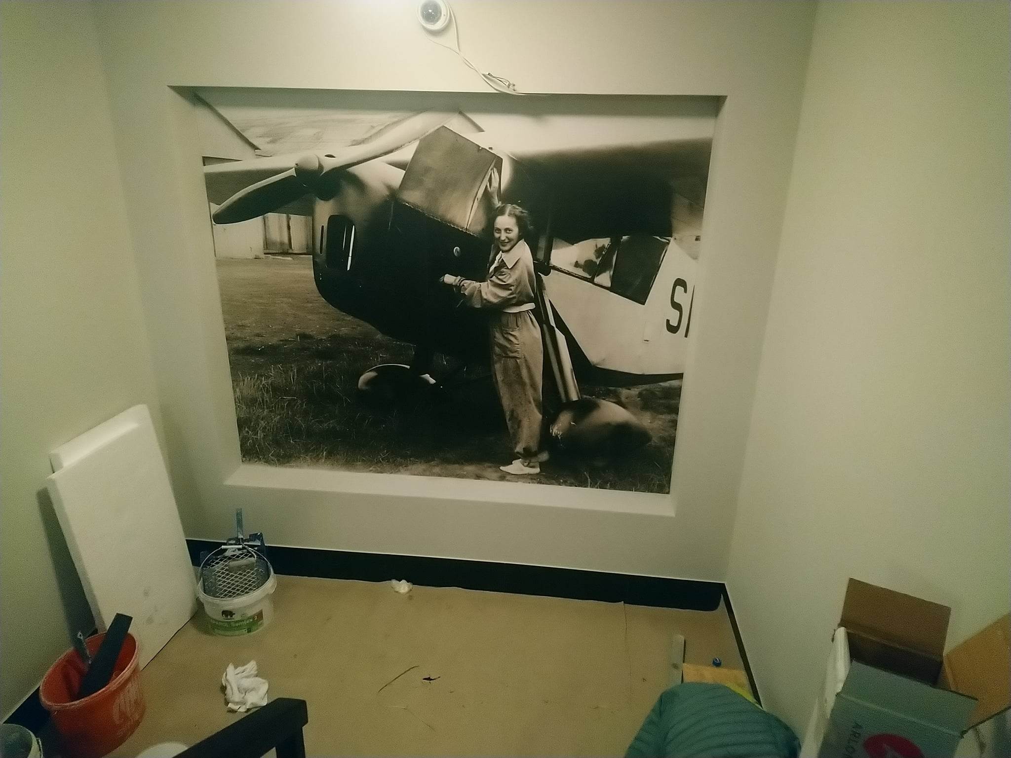 Czarno-biała fototapeta z wizerunkiem kobiety przy samolocie, zamontowana we wnęce ściany, z widocznymi narzędziami malarskimi i materiałami budowlanymi na podłodze.