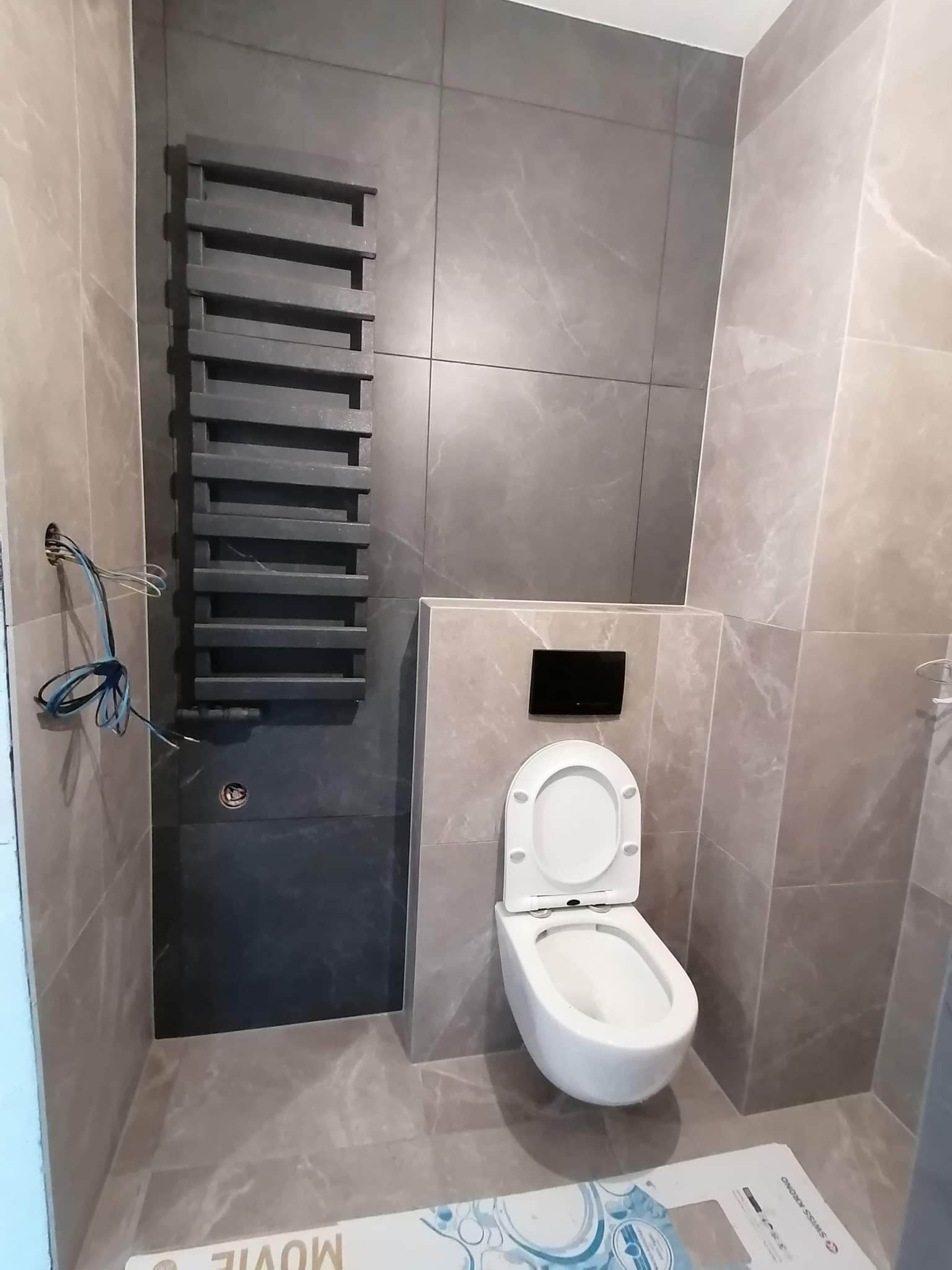 Wykończona łazienka z szarymi płytkami imitującymi marmur, wisząca toaleta z czarnym panelem spłukującym i drabinkowy grzejnik w kolorze grafitowym, z widocznymi przewodami elektrycznymi po lewej...