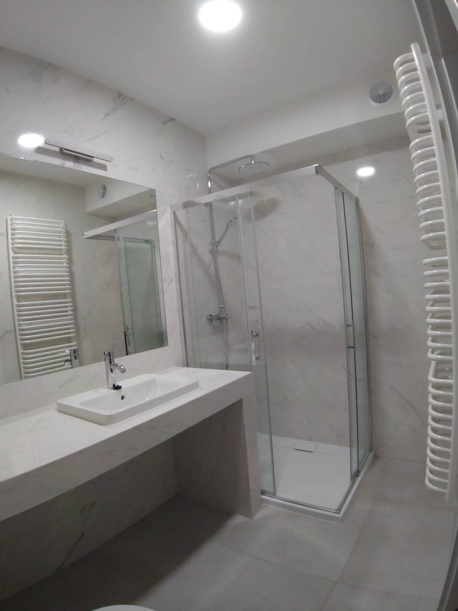 Nowoczesna łazienka z marmurowymi płytkami, przeszklona kabina prysznicowa typu walk-in z deszczownicą, biała umywalka nablatowa z baterią oraz grzejnik drabinkowy.