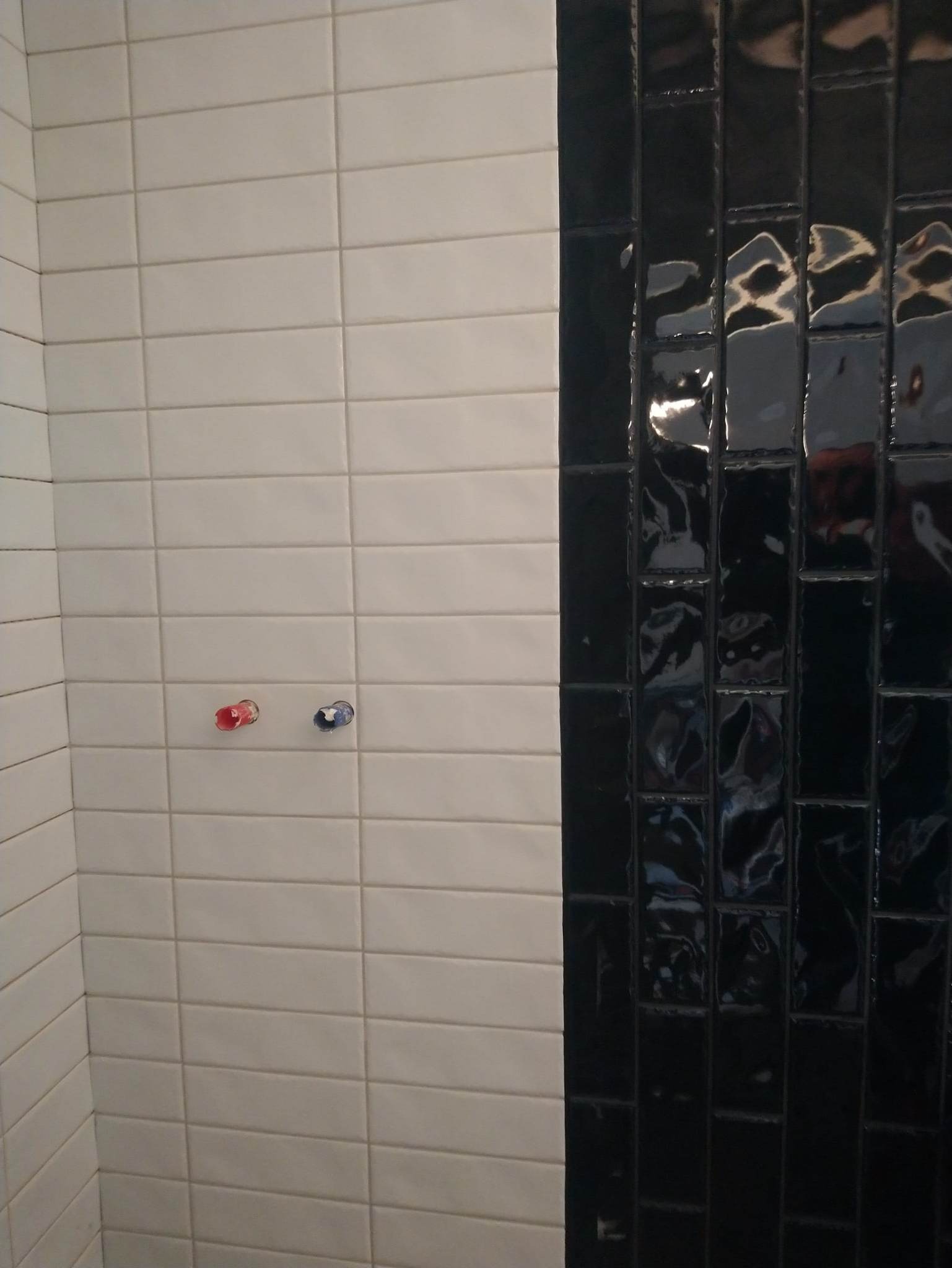 Ściana wyłożona białymi prostokątnymi płytkami z widocznymi końcówkami instalacji wodnej oraz pionowym pasem błyszczących, czarnych płytek.