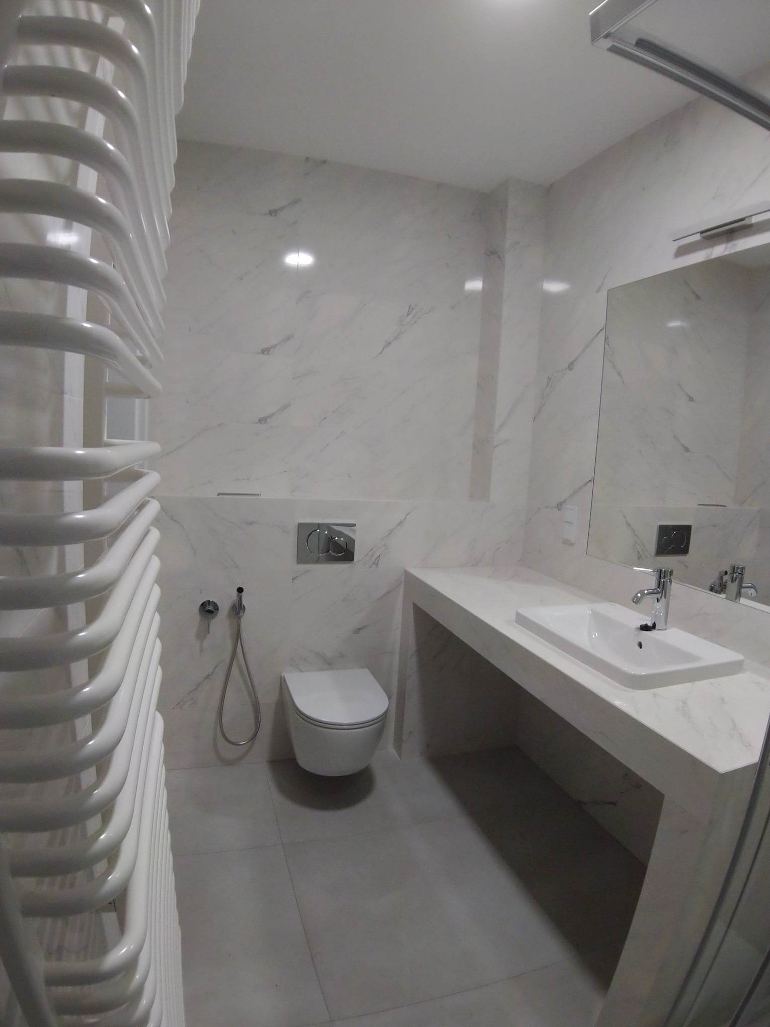 Nowoczesna, wykończona łazienka w Krakowie z białymi marmurowymi płytkami, podwieszaną toaletą, umywalką nablatową i grzejnikiem drabinkowym.