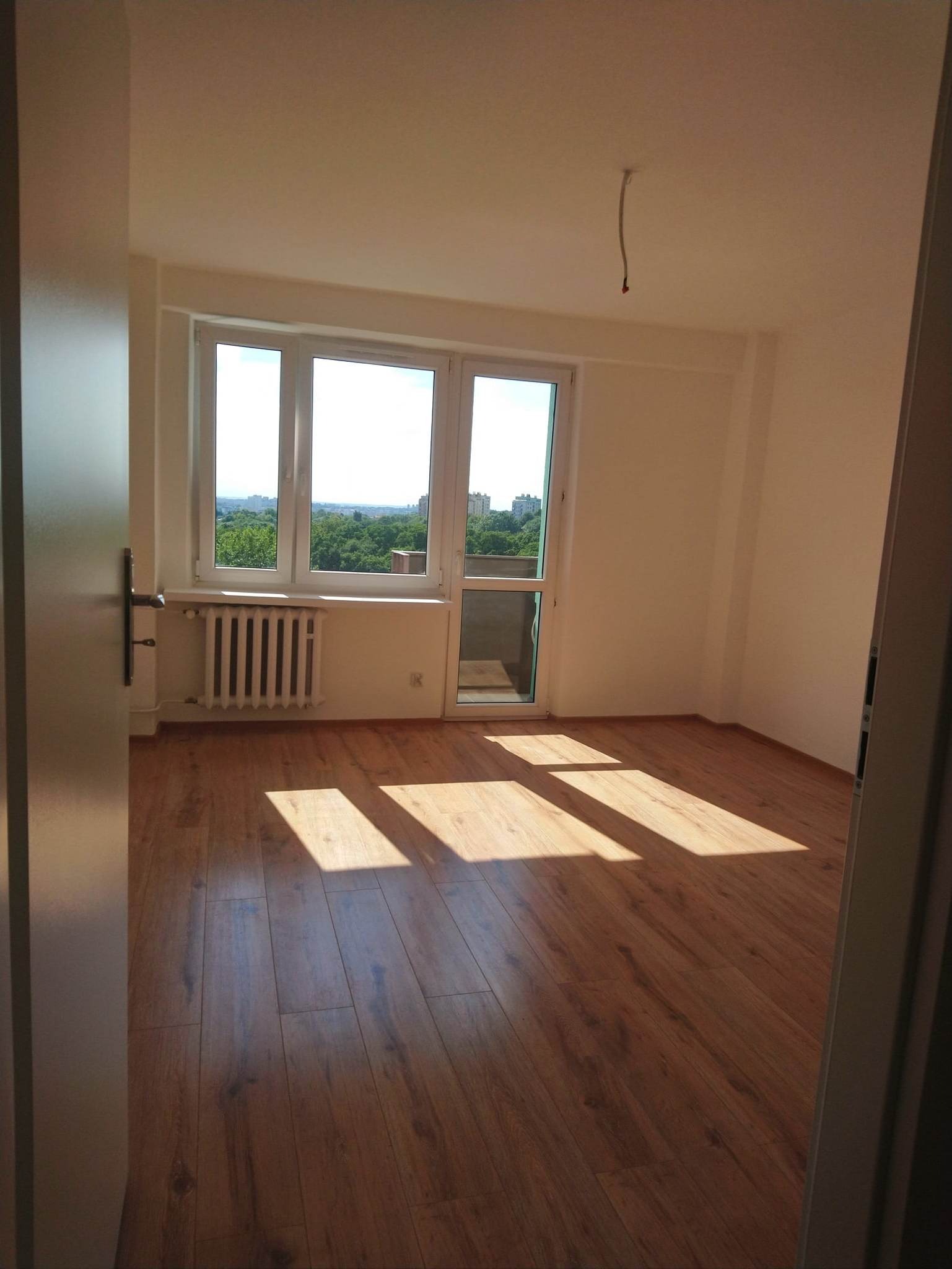 Pusty pokój po remoncie z drewnianą podłogą, świeżo pomalowanymi ścianami i dużym oknem z widokiem na zieleń miejską, przez które wpadają promienie słoneczne. Z sufitu wystaje kabel.