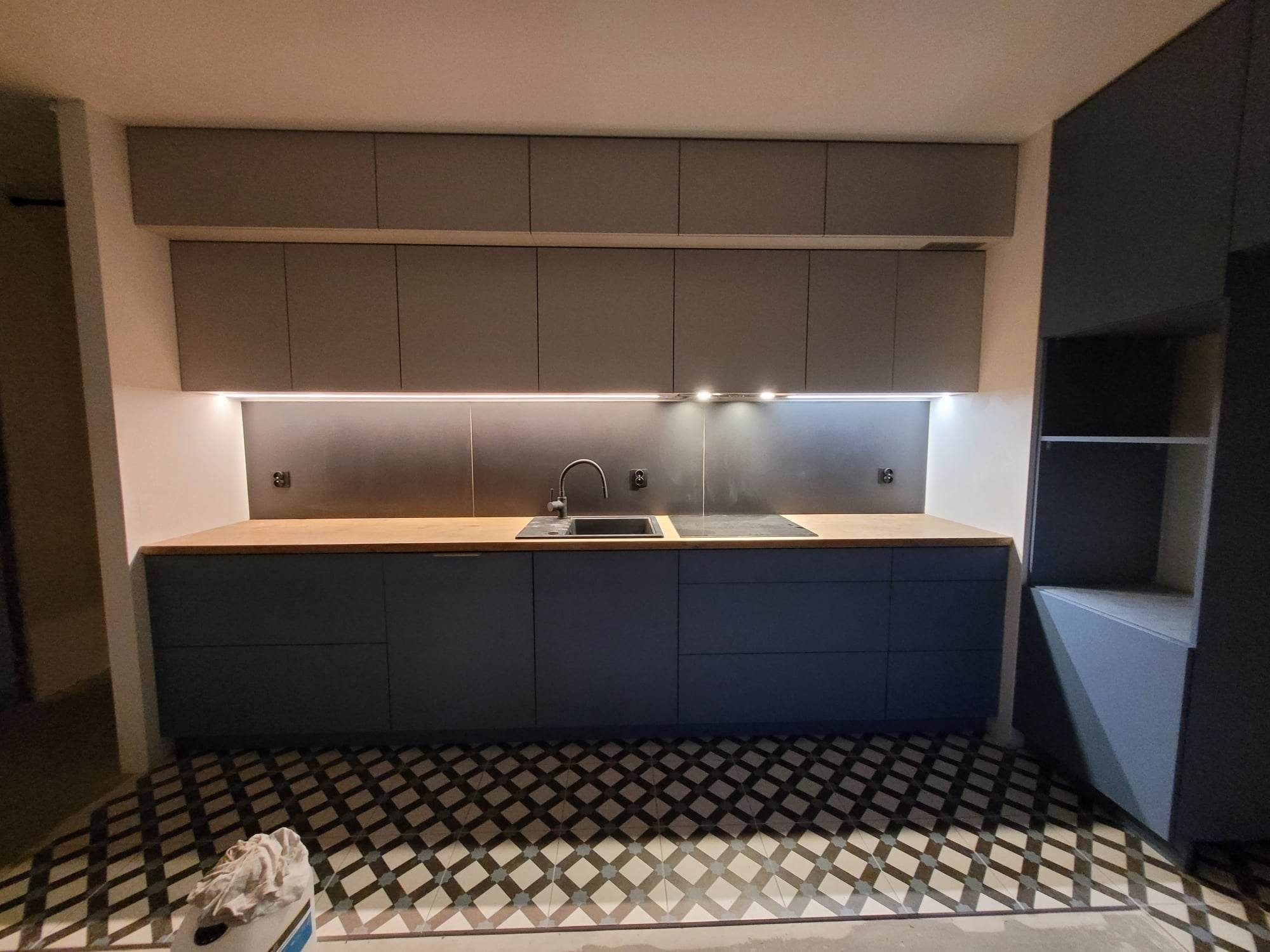 Nowoczesna kuchnia z ciemnymi, matowymi szafkami dolnymi i jasnoszarymi górnymi, drewnianym blatem, czarną armaturą i geometryczną podłogą w biało-czarną szachownicę. Widoczne oświetlenie LED...