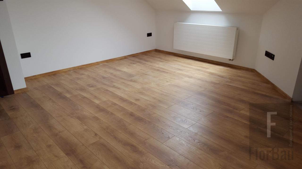 Jasny pokój na poddaszu z nowo ułożonymi panelami podłogowymi w odcieniach brązu i beżu, widoczne gniazdka elektryczne i biały grzejnik pod oknem dachowym.