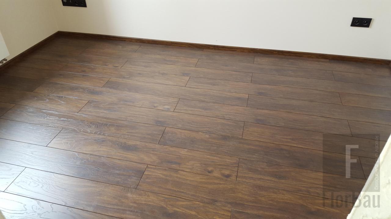 Ułożona podłoga z ciemnych paneli z widoczną strukturą drewna, brązowe listwy przypodłogowe, białe ściany z gniazdkami elektrycznymi.