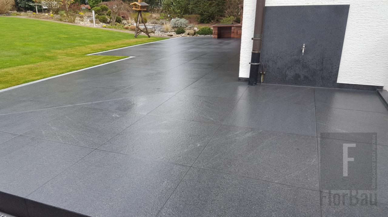 Nowoczesny taras z ciemnoszarych kamiennych płyt, widok na trawnik i elementy architektury ogrodowej w tle, lekko mokra powierzchnia.