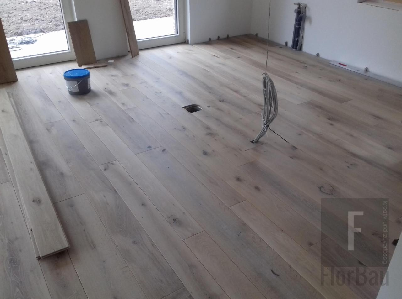 Jasna, drewniana podłoga z widocznymi przewodami wystającymi z otworu w podłodze, obok leży nieużywana klepka, a w tle widoczne okno i puszka z klejem.