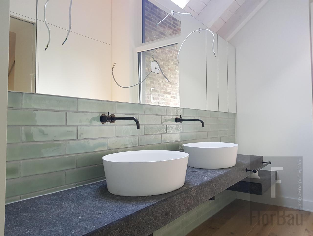 Nowoczesna łazienka z dwoma okrągłymi umywalkami nablatowymi, czarnymi bateriami i błyszczącymi, zielonymi płytkami ułożonymi w cegiełkę, widoczne kable do oświetlenia nad lustrem.