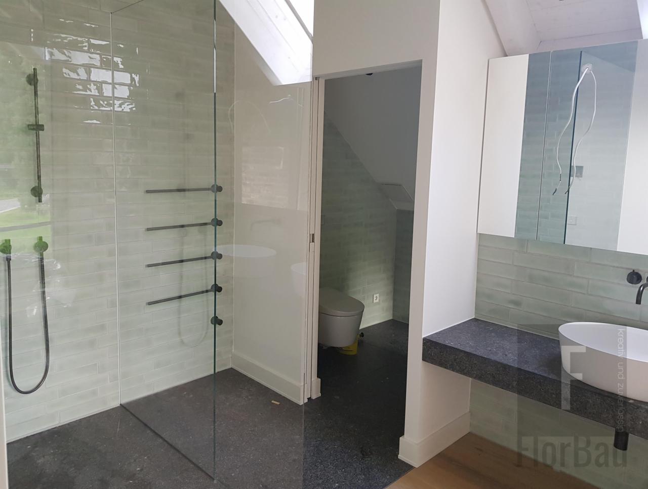 Nowoczesna łazienka z kabiną prysznicową typu walk-in, jasnymi, podłużnymi płytkami na ścianach, czarną armaturą i blatem z ciemnego kamienia, widok na toaletę w oddzielnym pomieszczeniu.