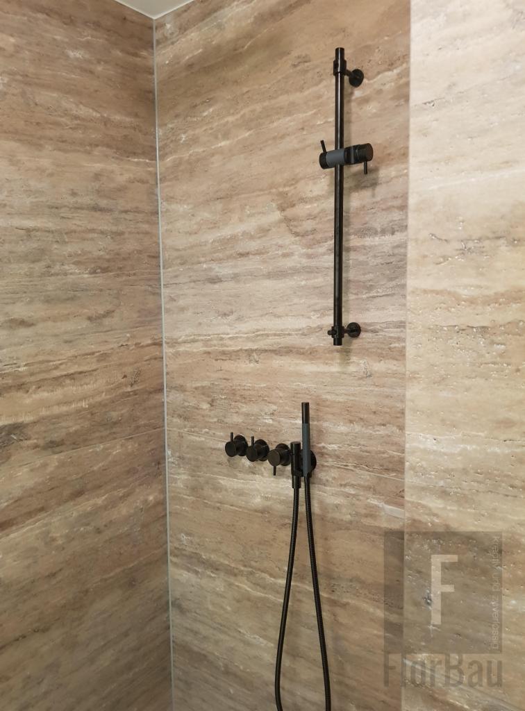 Wykończona łazienka z beżowymi płytkami imitującymi trawertyn, wyposażona w czarną armaturę: deszczownicę i baterię prysznicową z wężem. Widoczne delikatne logo firmy w prawym dolnym rogu.