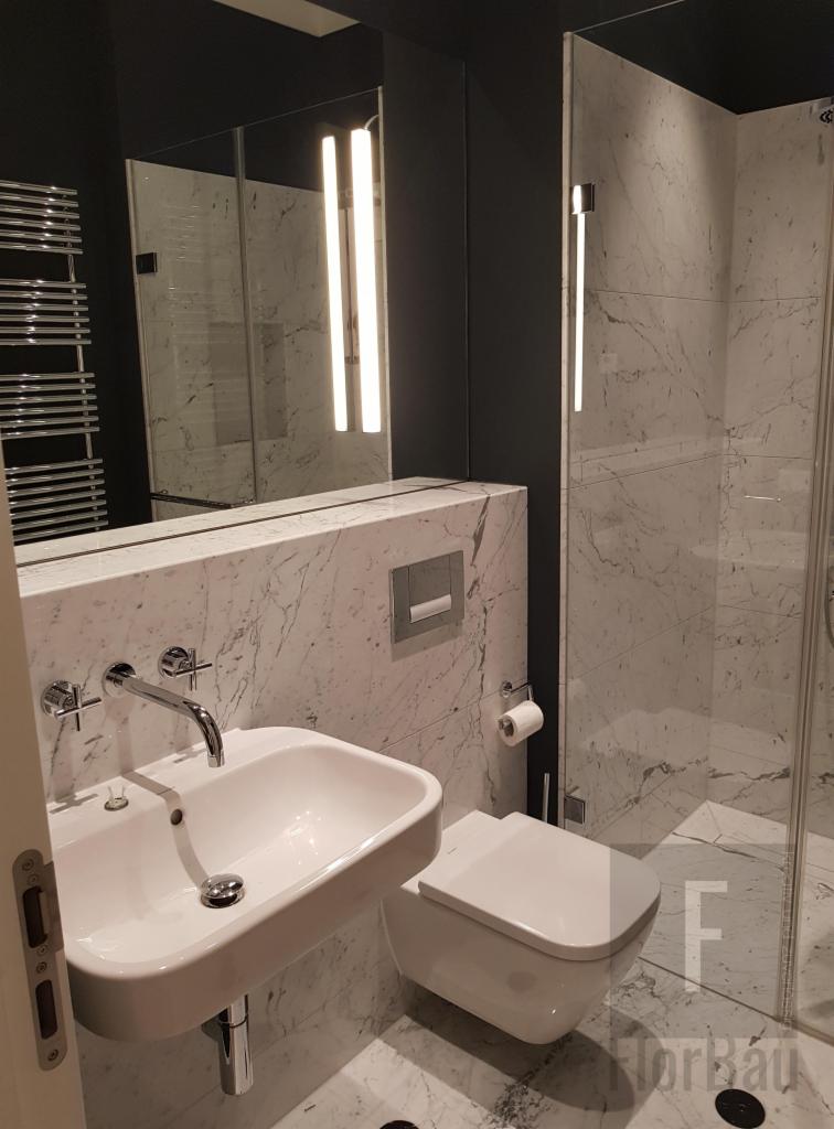 Nowoczesna łazienka z marmurowymi płytkami, białym umywalką i toaletą podwieszaną, oświetlonym lustrem i szklaną kabiną prysznicową.