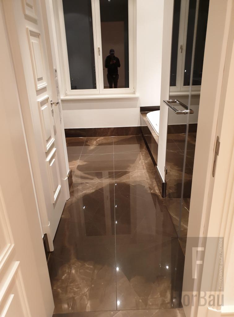 Błyszcząca, brązowo-marmurkowa podłoga w łazience z kabiną prysznicową ze szklanymi drzwiami i białymi ramami okiennymi, widok zza uchylonych, białych drzwi.