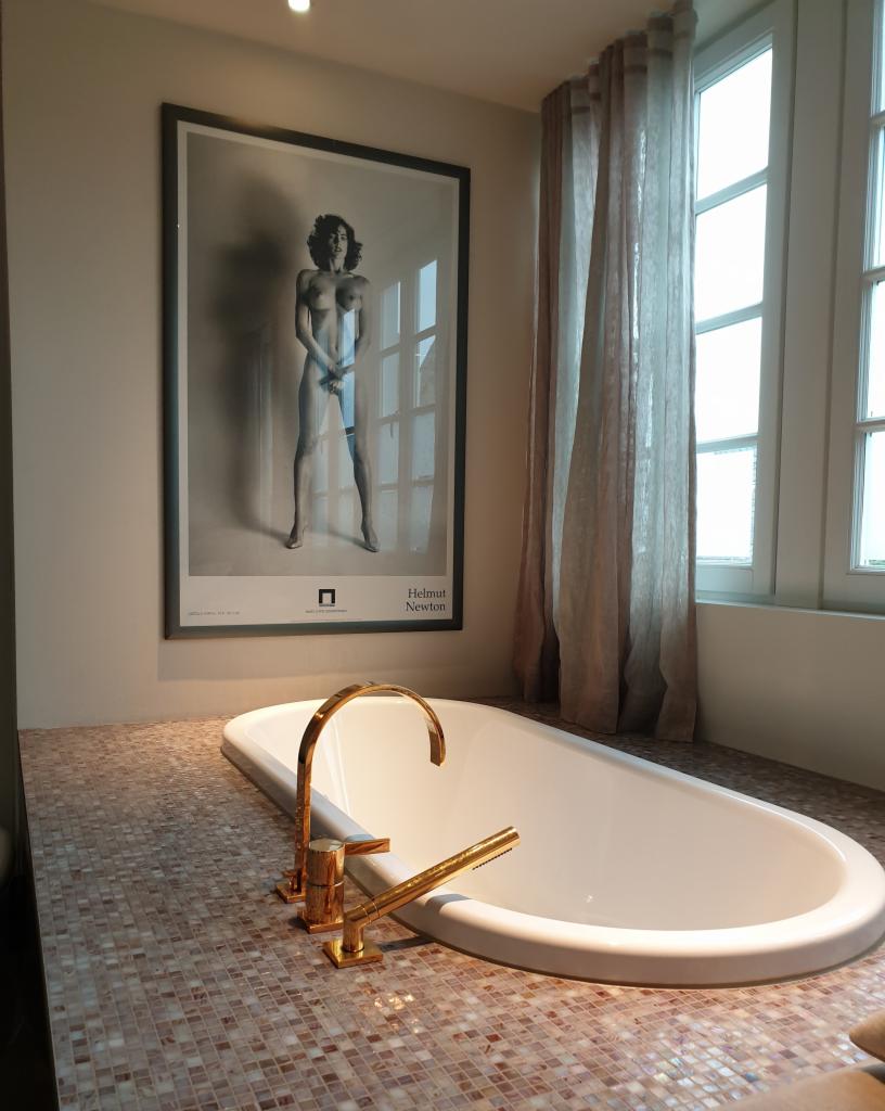 Biała wanna wolnostojąca z armaturą w kolorze złotym, otoczona mozaiką. Na ścianie wisi czarno-biały plakat w ramie. W tle okno z zasłonami.