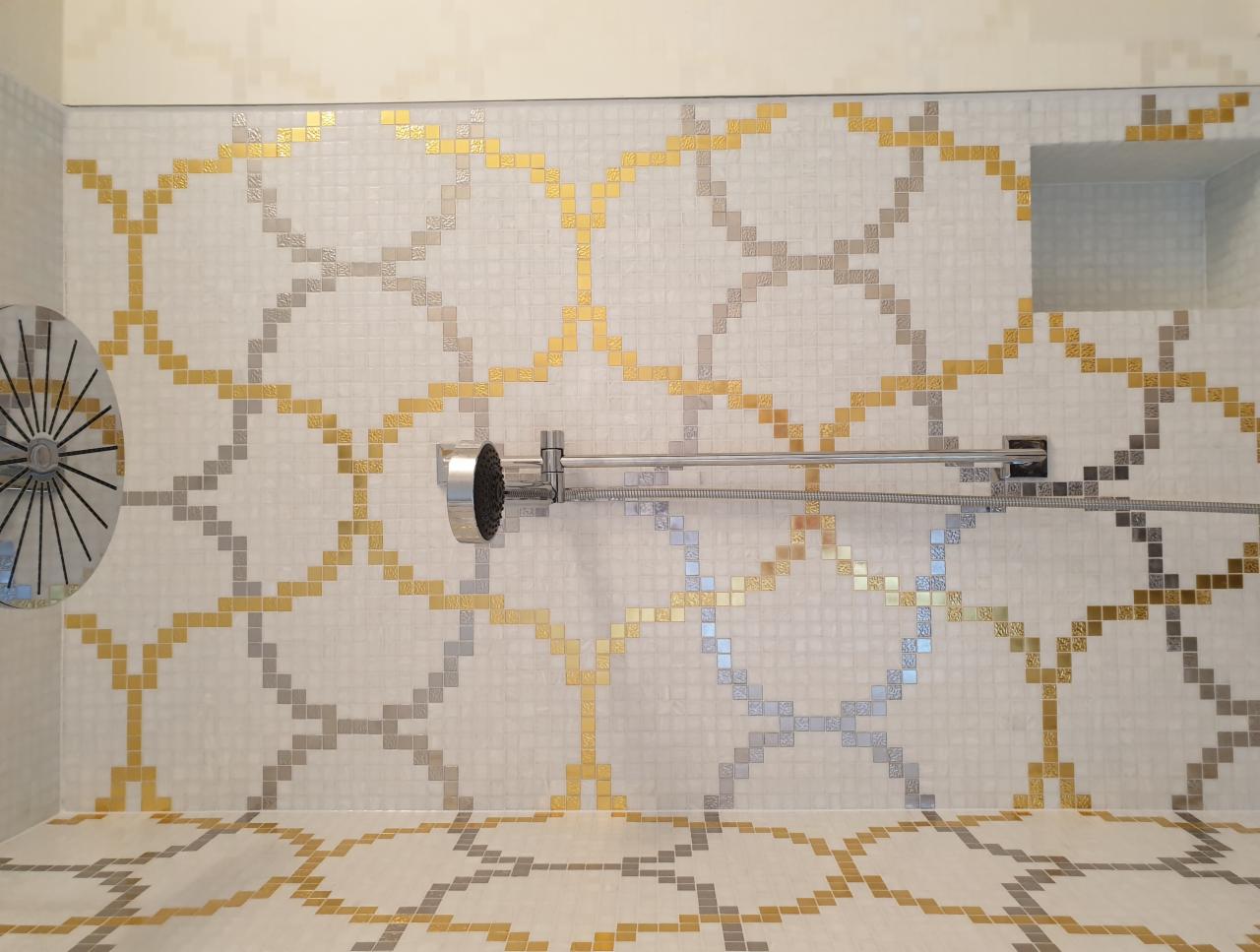 Kabina prysznicowa z mozaiką w odcieniach złota, srebra i bieli, tworzącą geometryczny wzór na ścianach i podłodze. Widoczna deszczownica i bateria prysznicowa.