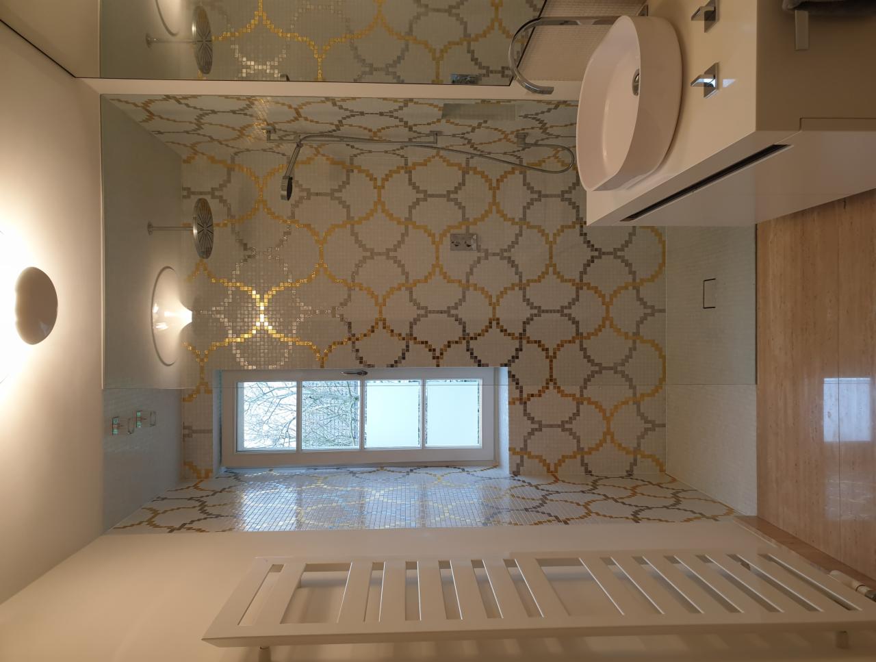 Nowoczesna łazienka z mozaiką w odcieniach bieli, szarości i złota, wzór geometryczny na ścianach i podłodze, biała umywalka nablatowa i grzejnik drabinkowy, okno z widokiem na drzewa.