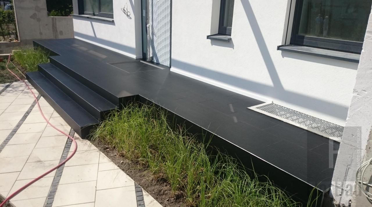 Nowoczesny taras z ciemnych płyt kamiennych z trzema stopniami, widok z boku, z niską roślinnością przy krawędzi i białym budynkiem w tle.