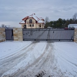 Metal-Spec - Czarna brama wjazdowa z pionowych profili, zamontowana na słupkach z piaskowca, prowadząca do domu z czerwoną dachówką, pokrytego śniegiem w zimowy dzień.