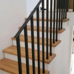 Schody drewniane z czarną, metalową balustradą o prostym, nowoczesnym designie, widoczne z boku, z białymi ścianami w tle.