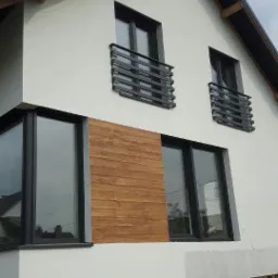 Nowoczesny dom z białym tynkiem, drewnianą okładziną, czarnymi oknami i minimalistycznymi, poziomymi balustradami balkonowymi, widoczne ocieplenie fundament&oacute;w i drabina.