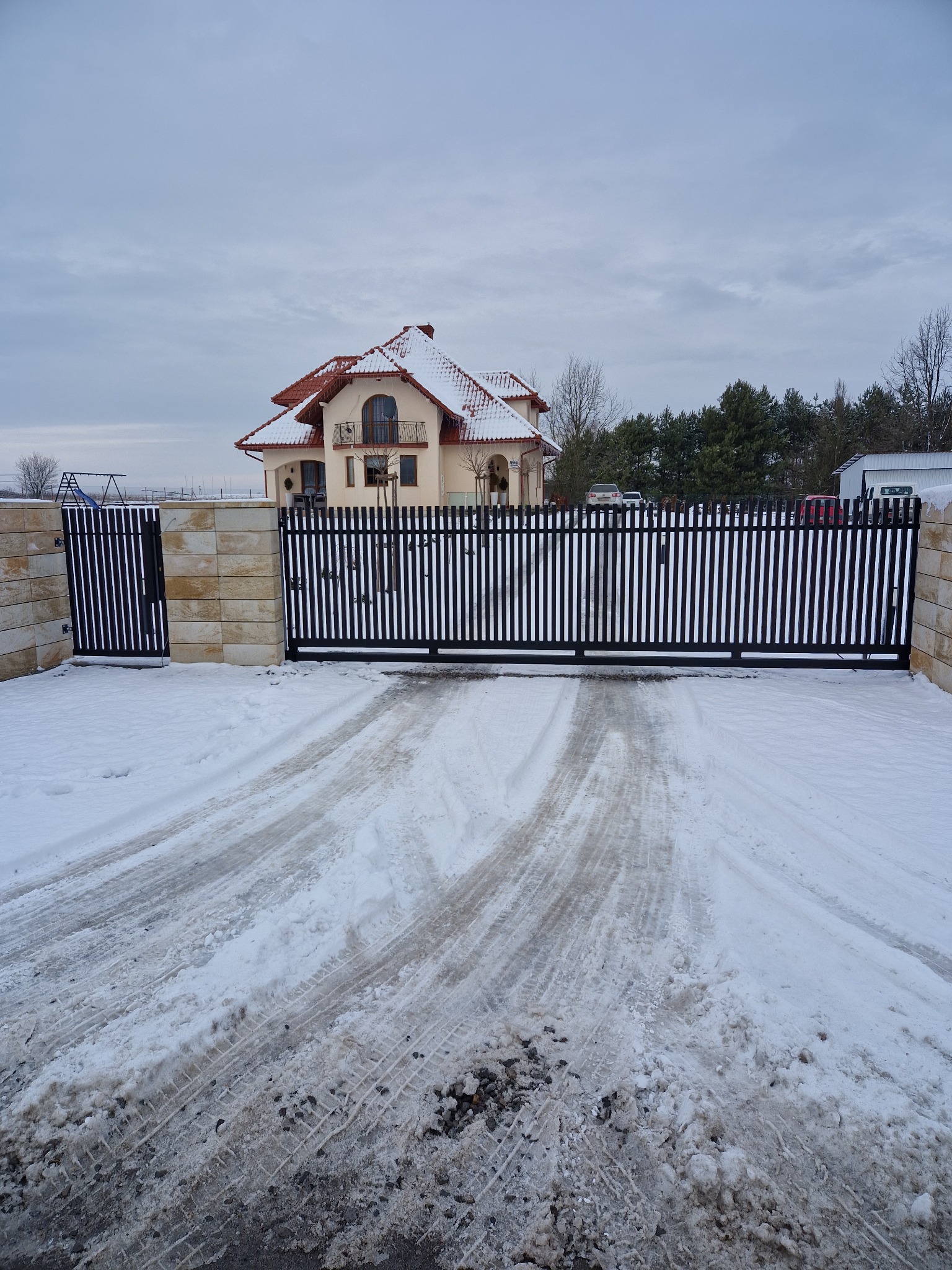 Czarna brama wjazdowa z pionowych profili, zamontowana na słupkach z piaskowca, prowadząca do domu z czerwoną dachówką, pokrytego śniegiem w zimowy dzień.