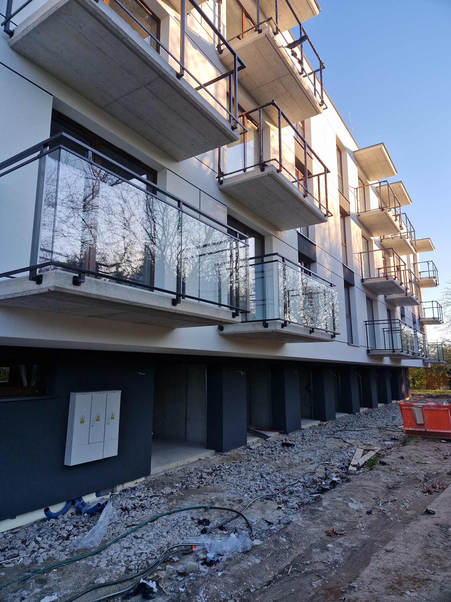 Nowoczesny budynek mieszkalny z balkonami z czarnymi ramami i szklanymi panelami, widok z dołu na tle błękitnego nieba, teren wokół budynku w trakcie prac wykończeniowych.