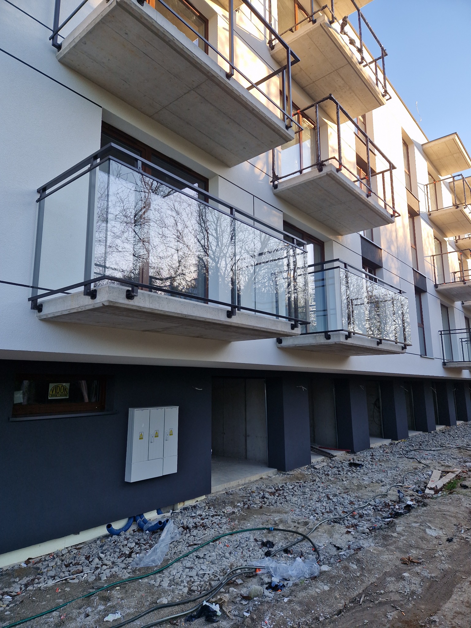 Nowoczesny budynek z balkonami z czarnymi ramami i szklanymi panelami, widok na plac budowy z gruzem i przewodami.