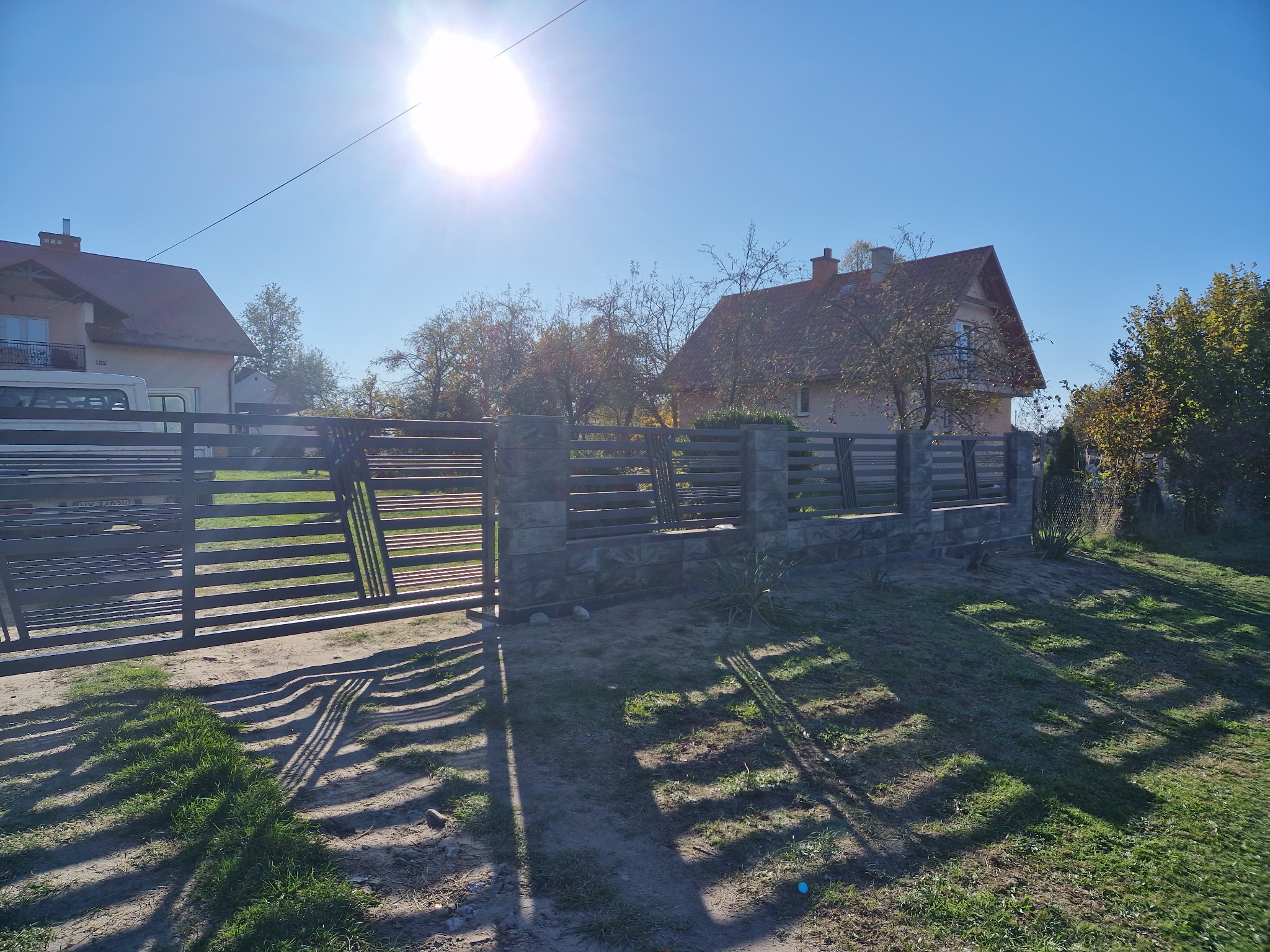 Nowoczesna brama wjazdowa z metalowymi przęsłami i kamiennymi słupkami w słoneczny dzień; w tle widoczne domy i drzewa.