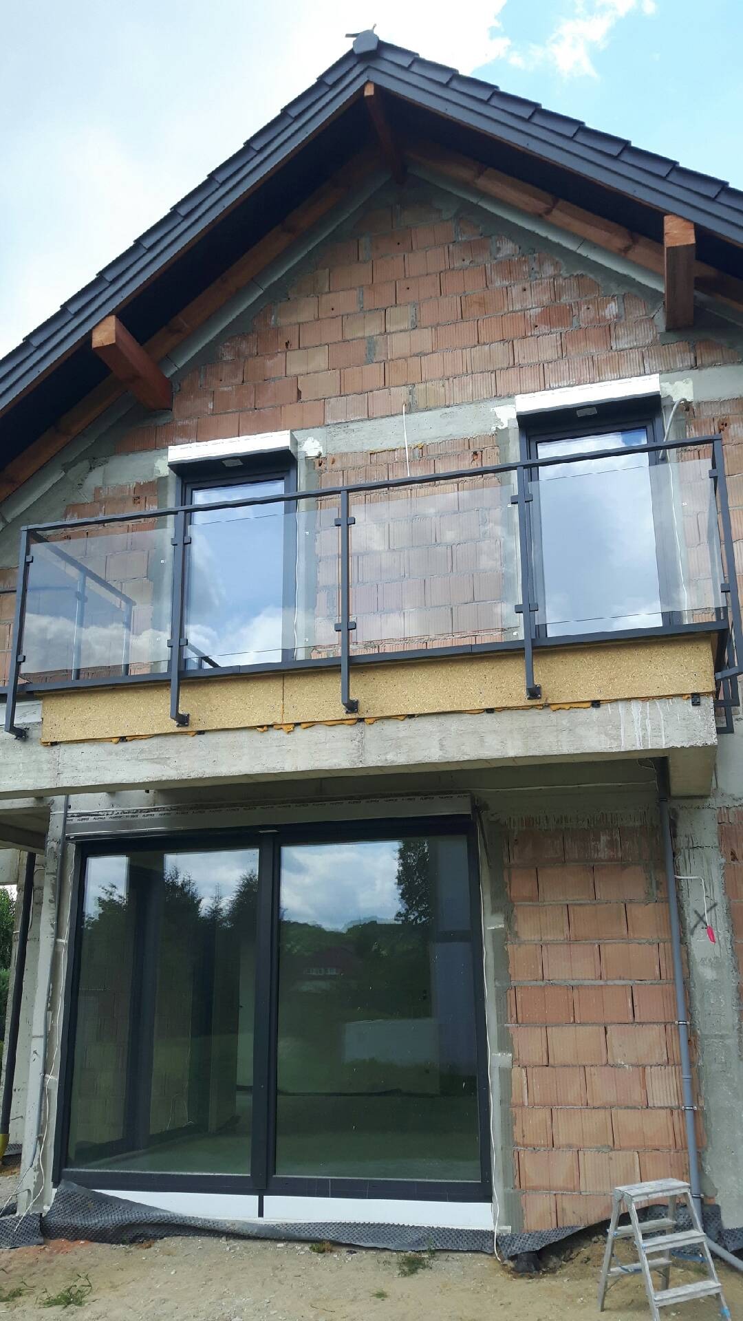 Balkon z czarną, metalową ramą i szklanym wypełnieniem, widoczny na tle surowej, ceglanej elewacji budynku w trakcie budowy, z widoczną drabiną i dużym oknem balkonowym.