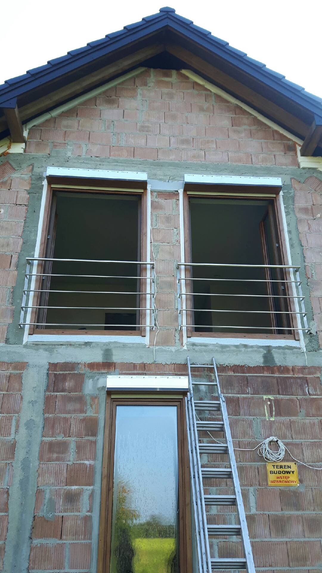 Surowe ściany budynku w trakcie budowy z zamontowanymi oknami i stalowymi balustradami balkonowymi, drabina oparta o ścianę i tabliczka informacyjna.