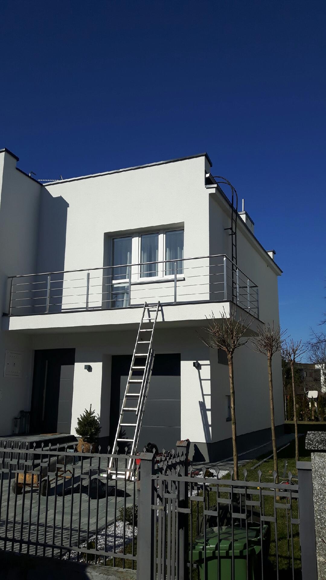 Nowoczesny dom z białymi ścianami i balkonem z poziomą balustradą, do którego oparta jest aluminiowa drabina. W tle błękitne niebo, a na pierwszym planie fragment ogrodzenia.